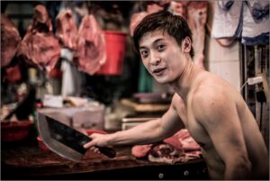 Young man with a cleaver in a butcher shop, shirtless and smiling.