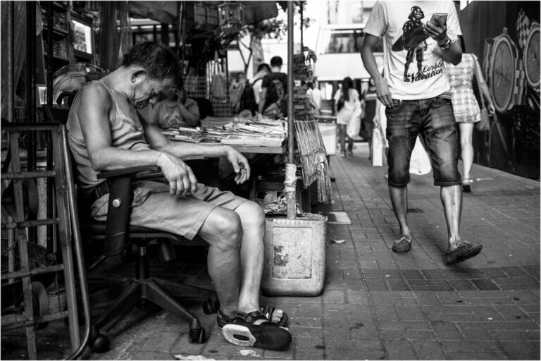 Pudsey Camera Club Member Image Black and white street scene with a man resting and a photographer walking by.